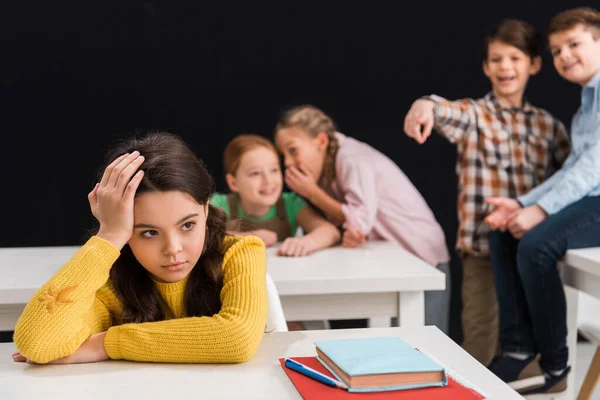 Enfoque selectivo de la colegiala frustrada cerca de compañeros de clase chismorreando mientras el escolar señala con el dedo aislado en negro, concepto de intimidación — Stock Photo