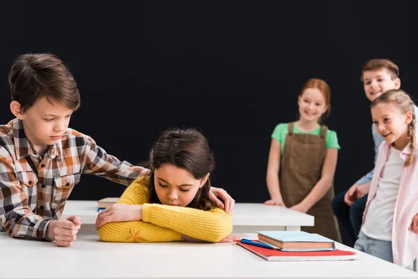 Selektiver Fokus des freundlichen Schülers, der aufgebrachte Schülerin in der Nähe lächelnder Klassenkameraden berührt, isoliert von schwarzem, Mobbing-Konzept — Stockfoto