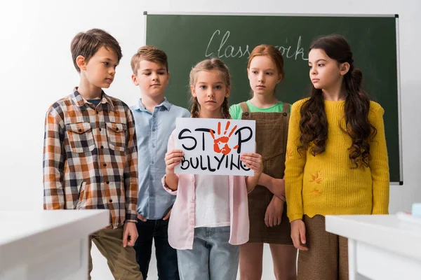 Enfoque selectivo de los escolares mirando a su compañero de clase con pancarta con letras de stop bullying - foto de stock