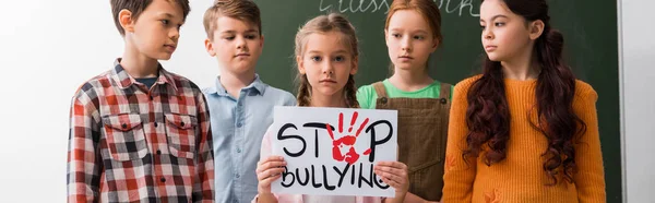 Plano panorámico de los escolares mirando a su compañero de clase con pancarta con letras de stop bullying - foto de stock