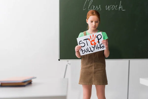Selective focus of sad kid holding placard with stop bullying lettering in classroom — Stock Photo