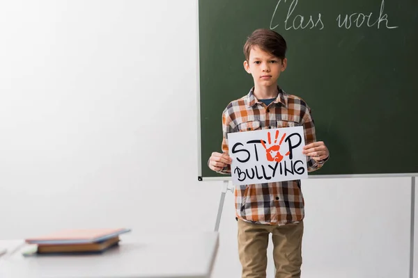 Selective focus of schoolboy holding placard with stop bullying lettering in classroom — Stock Photo
