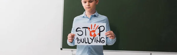 Panoramic shot of schoolboy holding placard with stop bullying lettering near chalkboard — Stock Photo