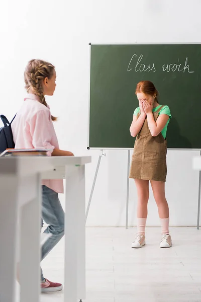 Enfoque selectivo de la colegiala mirando a su compañero de clase cubriendo la cara mientras llora en la escuela, concepto de intimidación - foto de stock