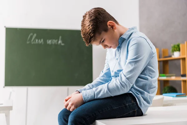 Sad and bullied schoolchild sitting on table in classroom — Stock Photo