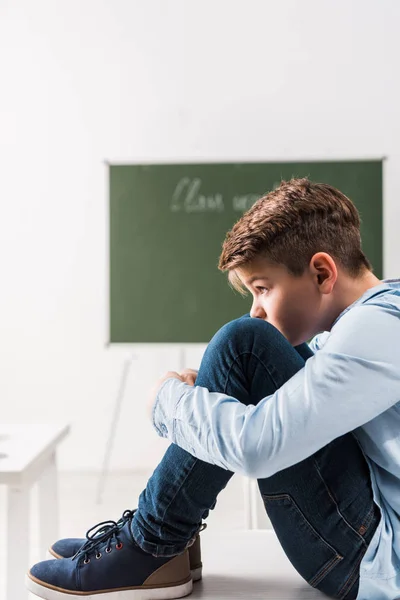 Bullied schoolboy sitting on table in classroom — Stock Photo