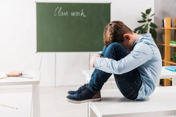 Escolar intimidado cubriendo la cara mientras llora y se sienta en la mesa - foto de stock