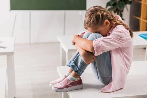 Gemobbtes Schulkind verdeckt Gesicht, während es auf Schreibtisch im Klassenzimmer sitzt — Stockfoto