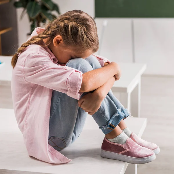 Bullied schoolgirl covering face while sitting on desk in classroom — Stock Photo