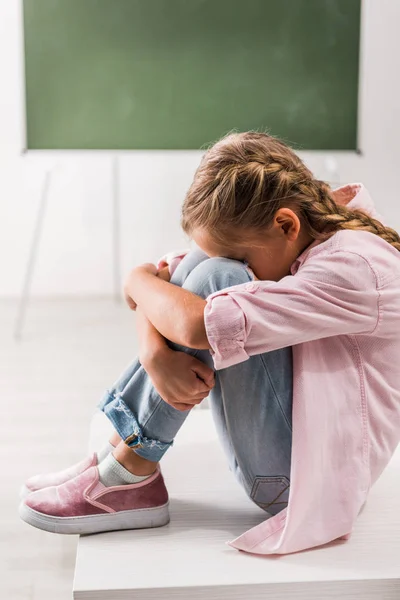 Molesto y acosado colegiala cubriendo la cara mientras está sentado en el escritorio en el aula - foto de stock