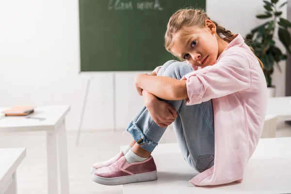 Bullied schoolgirl sitting on desk and looking at camera in classroom — Stock Photo