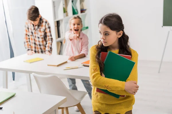 Selective focus of bullied schoolgirl holding notebooks near cruel classmate pointing with finger — Stock Photo