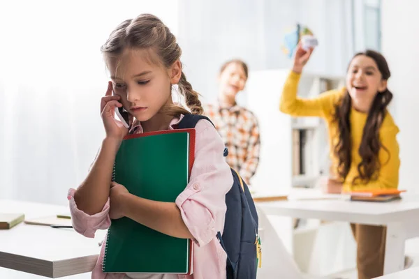 Selective focus of bullied schoolgirl talking on smartphone near cruel schoolkids in classroom — Stock Photo