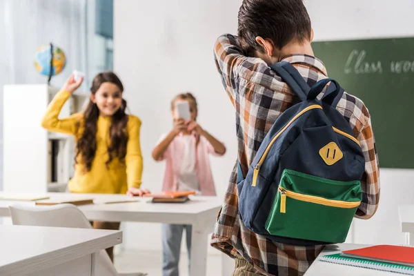 Vista posterior del niño acosado de pie cerca de compañeros de clase crueles con teléfono inteligente, concepto de acoso cibernético - foto de stock