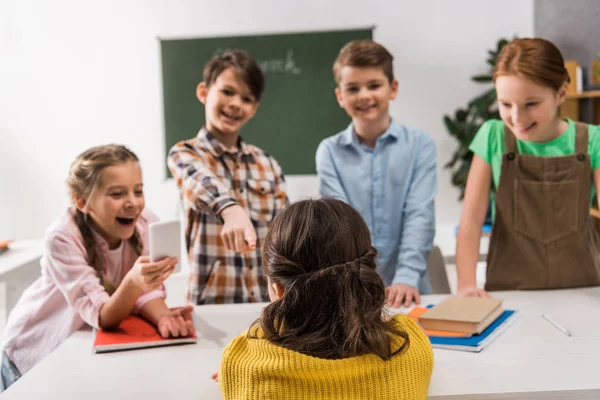 Rückansicht gemobbte Schülerin sitzt neben Schulkind mit Smartphone und grausamen Klassenkameraden, Konzept Cybermobbing — Stockfoto