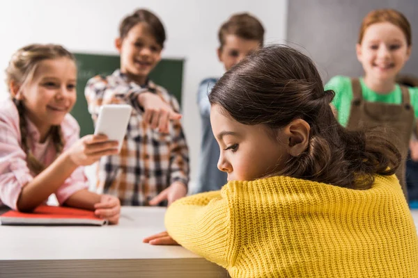 Focus selettivo della studentessa vittima di bullismo seduta vicino a scuola con smartphone e compagni di classe crudeli, concetto di cyberbullismo — Foto stock