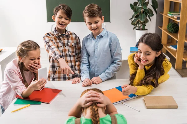 Focus selettivo dello scolaro con smartphone e compagni di classe crudeli che ridono vicino al bambino vittima di bullismo, concetto di cyberbullismo — Foto stock