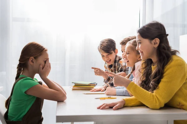 Colegiales crueles señalando con los dedos y riendo cerca de la colegiala acosada cubriendo la cara mientras llora en el aula - foto de stock