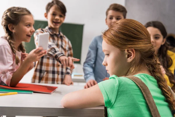 Selective focus of depressed schoolgirl sitting near schoolkid with smartphone and cruel classmates, cyberbullying concept — Stock Photo