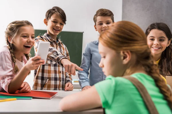 Focus selettivo di scolaro con smartphone e compagni di classe crudeli che ridono vicino al bambino vittima di bullismo, concetto di cyberbullismo — Foto stock