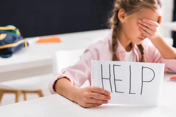 Enfoque selectivo de papel con letras de ayuda en la mano de la colegiala molesta cubriendo los ojos, concepto de intimidación - foto de stock