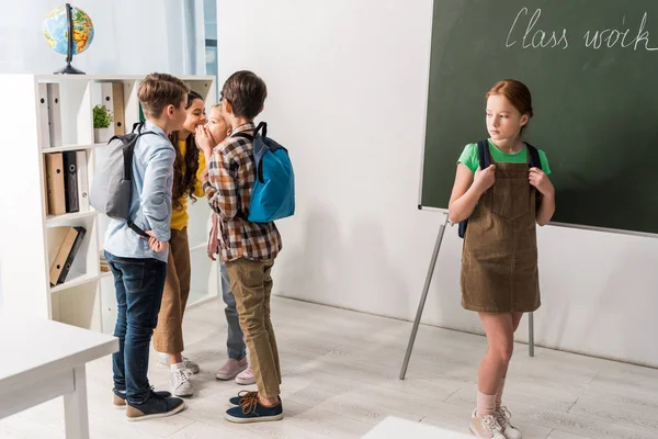 Cruels écoliers bavardage et rire près intimidé écolière debout dans la salle de classe — Photo de stock