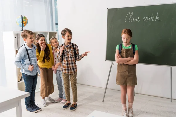 Cruel colegial señalando con el dedo y riendo cerca de colegiala intimidado de pie en el aula - foto de stock