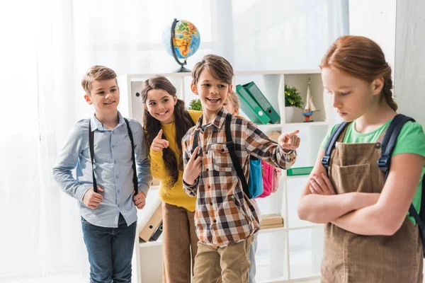 Enfoque selectivo de escolares crueles señalando con los dedos y riendo cerca de colegiala intimidada de pie con los brazos cruzados en el aula - foto de stock