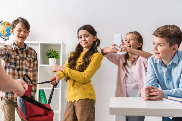 Selective focus of cruel schoolkid using smartphone near classmates while bullying schoolgirl — Stock Photo