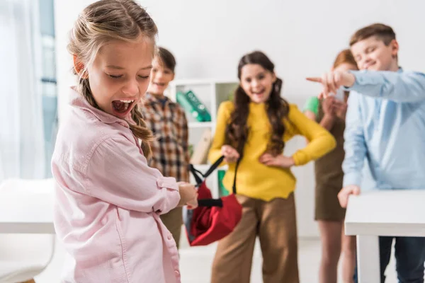 Enfoque selectivo de la colegiala acosada gritando cerca de crueles pupilas crueles sosteniendo la mochila y señalando con los dedos - foto de stock