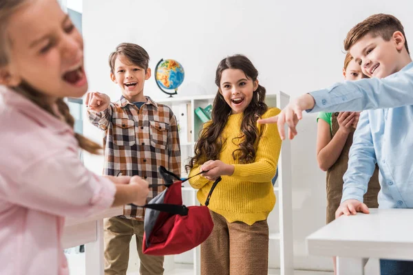 Enfoque selectivo de alumnos crueles sosteniendo la mochila y señalando con los dedos a su compañero de clase acosado gritando en el aula - foto de stock