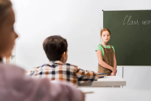 Enfoque selectivo de la colegiala molesta de pie cerca de pizarra y compañeros de clase - foto de stock