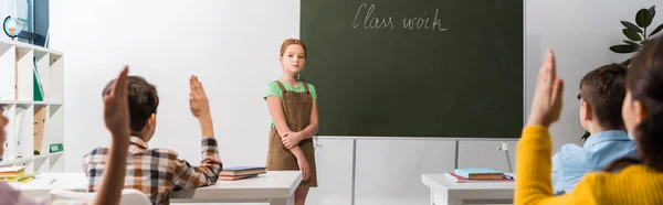 Panoramic shot of schoolkgirl standing near chalkboard with class work lettering and classmates with raised hands — Stock Photo