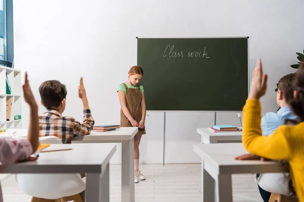 Messa a fuoco selettiva di scolaro in piedi vicino alla lavagna con lettere di lavoro di classe e compagni di classe con le mani alzate — Foto stock