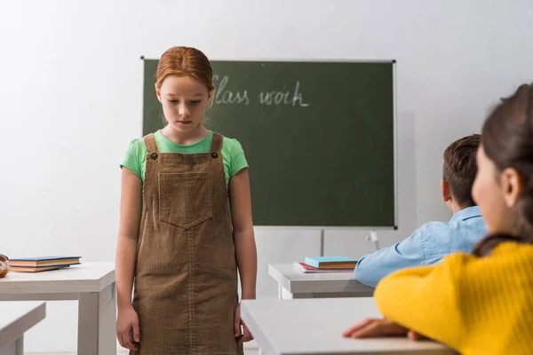 Enfoque selectivo de la colegiala molesta caminando cerca de compañeros de clase sentados en escritorios - foto de stock