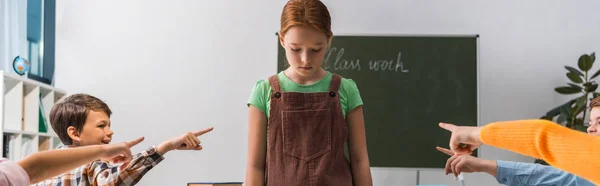 Panoramic shot of cruel classmates pointing with fingers at depressed bullied schoolgirl in classroom — Stock Photo