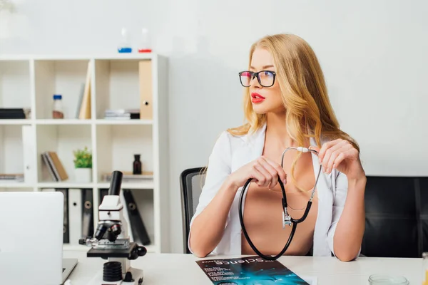 Sexy nurse in glasses holding stethoscope near microscope in laboratory — Stock Photo