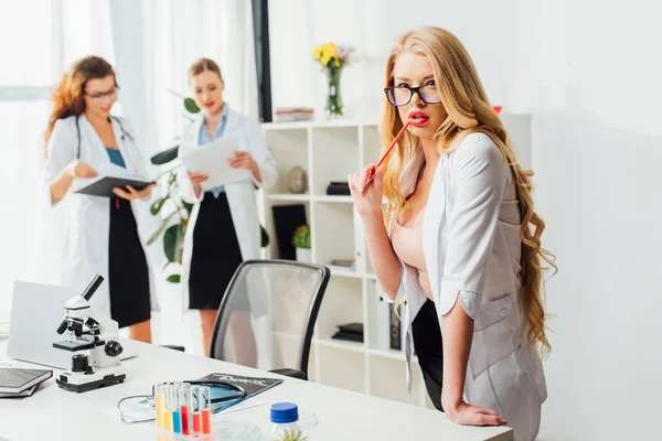 Selective focus of sexy nurse holding pencil near lips and standing with women in laboratory — Stock Photo