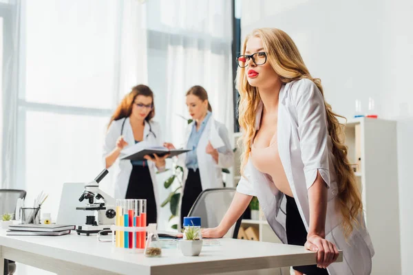 Selektiver Fokus der sexy Krankenschwester mit Brille und weißem Mantel in der Nähe von Frauen im Labor — Stockfoto