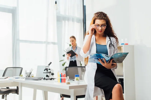 Selektiver Fokus der sexy Krankenschwester in Brille und weißem Mantel beim Lesen eines Wissenschaftsmagazins nahe der Frau im Labor — Stockfoto