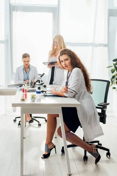 Sexy nurse holding glasses while sitting near women in white coats in laboratory — Stock Photo