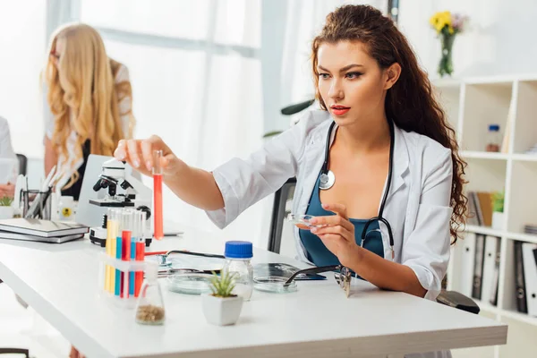 Infirmière sexy tenant tube à essai tout en étant assis près de la femme en manteau blanc en laboratoire — Photo de stock