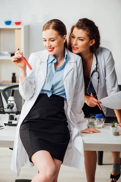 Счастливые и сексуальные медсестры смотрят на пробирку с красной жидкостью в лаборатории — стоковое фото