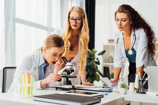 Enfoque selectivo de la enfermera sexy mirando a través del microscopio cerca de las mujeres - foto de stock