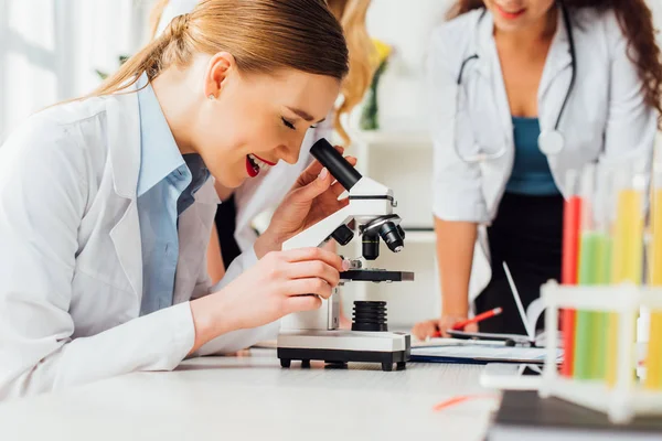 Selective focus of happy girl looking through microscope near sexy nurses — Stock Photo