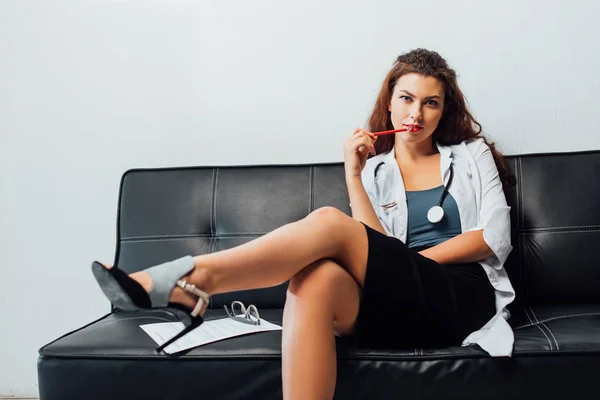 Sexy nurse sitting on sofa and holding pencil near lips — Stock Photo