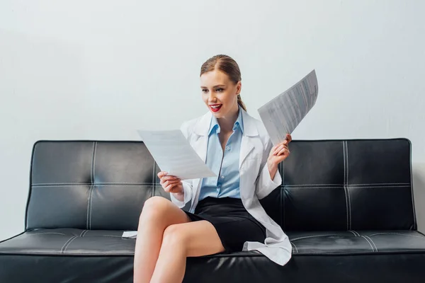 Enfermera feliz y sexy sentada en el sofá y sosteniendo papel - foto de stock