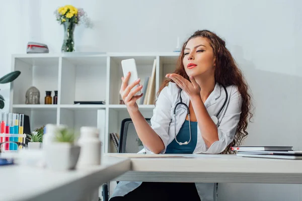 Selective focus of curly and sexy nurse sending air kiss while taking selfie in clinic — Stock Photo