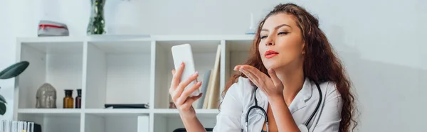 Panoramic shot of curly and sexy nurse sending air kiss while taking selfie in clinic — Stock Photo