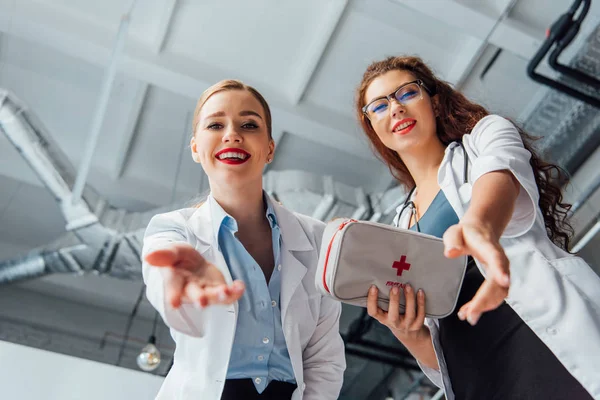 Tiefansicht glücklicher und sexy Krankenschwestern in weißen Mänteln mit Verbandskasten und ausgestreckten Händen — Stockfoto